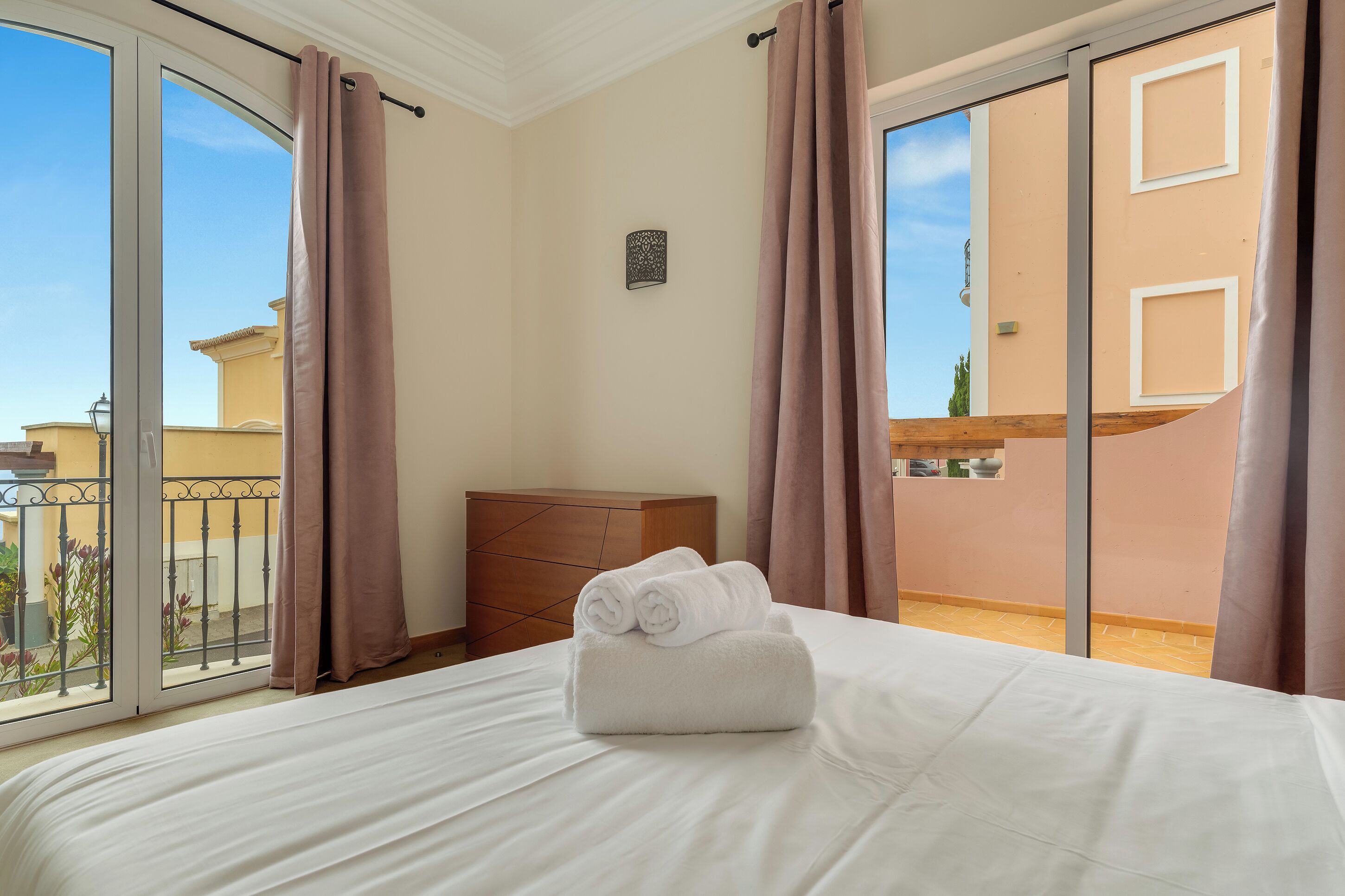 A hotel room with a bed, a wooden dresser, and a balcony with curtains.