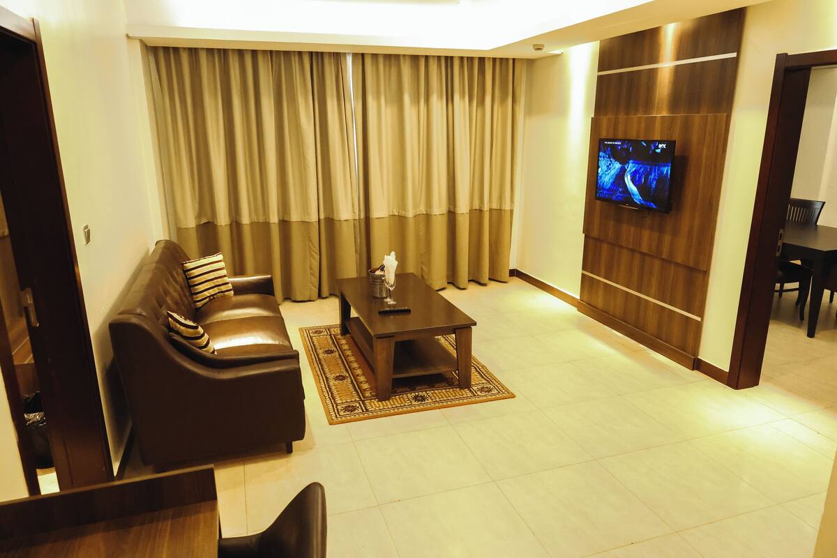 A living room with a brown leather sofa, a wooden coffee table, a TV mounted on a wooden wall, and a large window with beige curtains.
