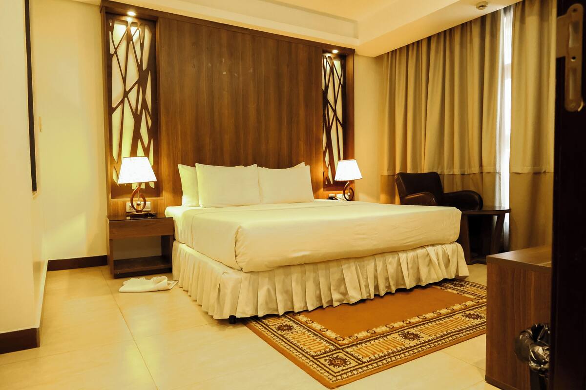 A hotel room with a large bed, two bedside tables with lamps, a wooden headboard, a brown rug, and a dark-colored desk.