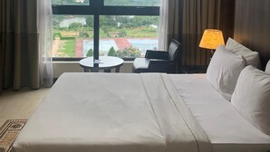 A hotel room with a large bed, a desk, and a chair. There is a view of a green landscape through the window. - Agulu Lake Resort and Conference Centre (Adazi Nnukwu)