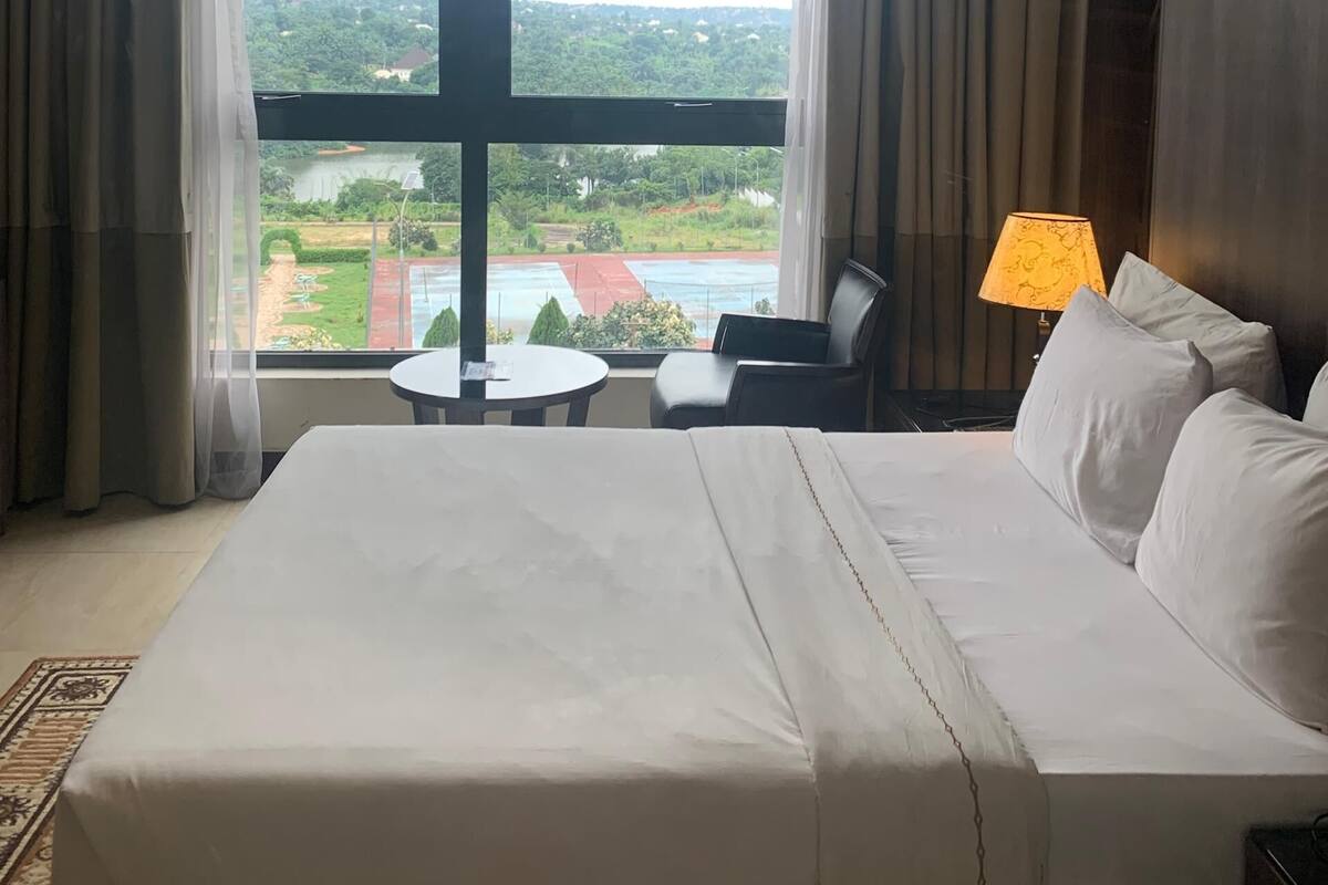 A hotel room with a large bed, a desk, and a chair. There is a view of a green landscape through the window.