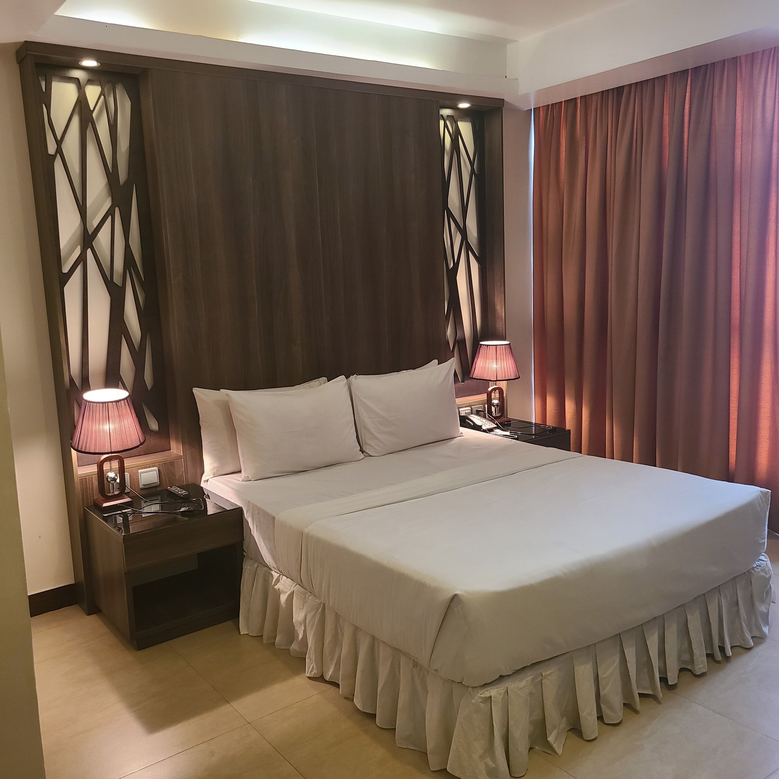 A large bed with a ruffled skirt, two bedside tables with lamps, a wooden headboard with a geometric design, and a curtain on the right.