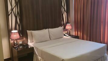 A large bed with a ruffled skirt, two bedside tables with lamps, a wooden headboard with a geometric design, and a curtain on the right.