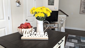 A wooden table with a 'Welcome' sign, a bowl of red apples, and a vase of yellow flowers.
