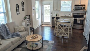 A living room with a sofa, coffee table, and a kitchen area with a dining table, chairs, and appliances. - Tiny Cottage w/private workspace in the heart of Davidson! (Davidson)