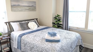 A neatly made bed with a floral-patterned bedspread, a large decorative pillow, and a folded towel on top. There is a bedside table with a lamp and a small plant. A painting hangs on the wall above the bed, and a window with curtains is visible.