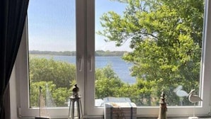 A room with a large window overlooking a lake, a radiator below, and a small table with decorative items. - Dream View Comfortable Holiday Residence (Maasholm)
