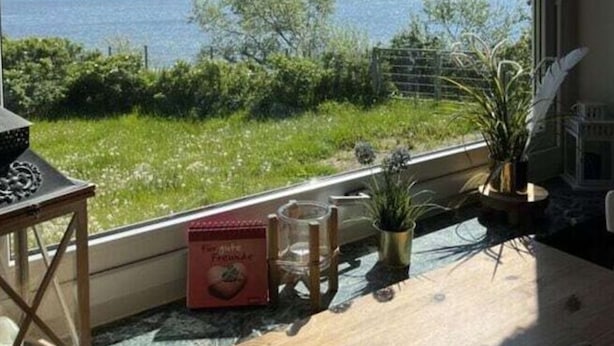 A room with a large window overlooking a body of water, a wooden table with a book and a plant, and a chair. - Dream View Comfortable Holiday Residence (Maasholm)