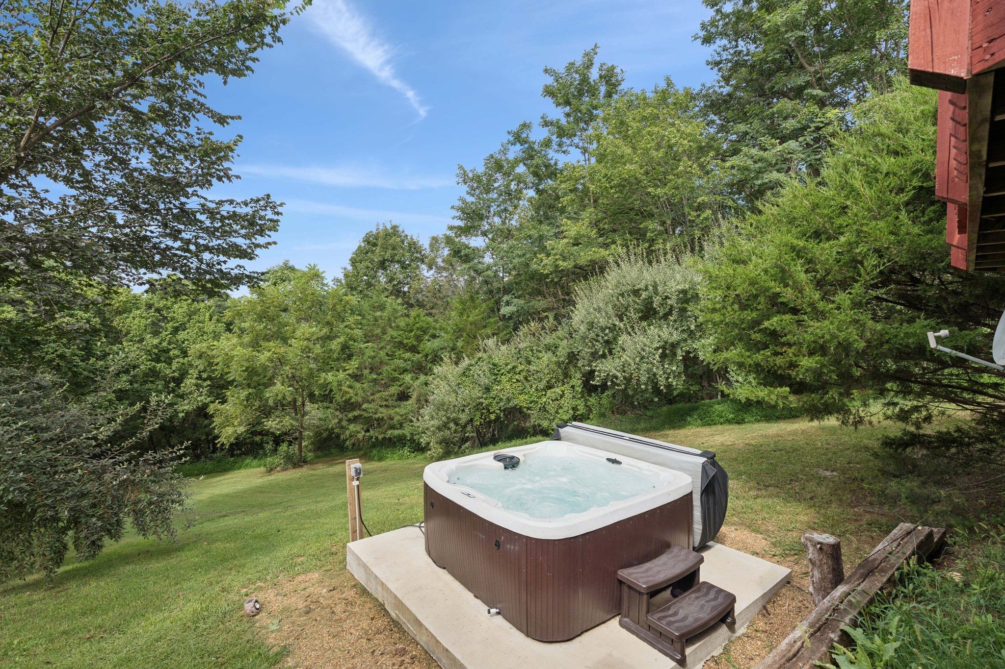 Un bain à remous dans un jardin, avec une terrasse en bois, entouré d’arbres et d’un bâtiment rouge.