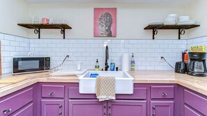 A kitchen with a microwave, sink, and cabinets. There are two shelves above the sink, one with dishes and the other with a painting.
