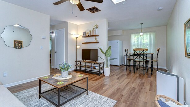 A spacious living room with a ceiling fan, a dining area with a table and chairs, a kitchen with a refrigerator, and a large window allowing natural light.