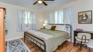 A bedroom with a bed, a desk, a chair, a ceiling fan, and a painting on the wall.