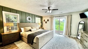 Ein Schlafzimmer mit einem großen Bett, einem Deckenventilator, einem Fenster mit Jalousien, einem Nachttisch, einer Kommode und Blick ins Freie.