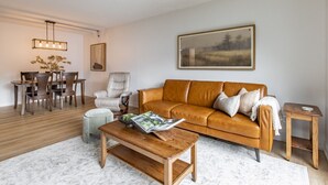 Un salon avec un canapé en cuir marron, une table basse en bois, une table à manger avec des chaises et un tableau accroché au mur.