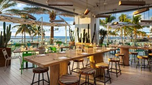 An outdoor bar area with wooden tables and stools, surrounded by potted plants and umbrellas, overlooking a beach with palm trees.