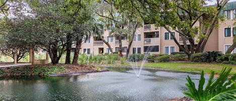 A pond with a fountain, surrounded by trees and a building with balconies.