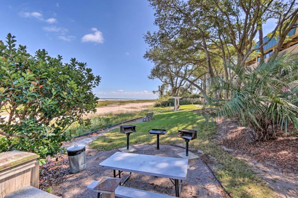 A patio area with a table and benches, surrounded by greenery and trees.