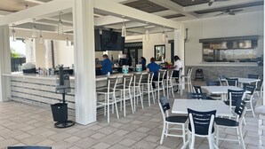 Un café al aire libre con mesas y sillas blancas, una zona de bar y una cocina al fondo.