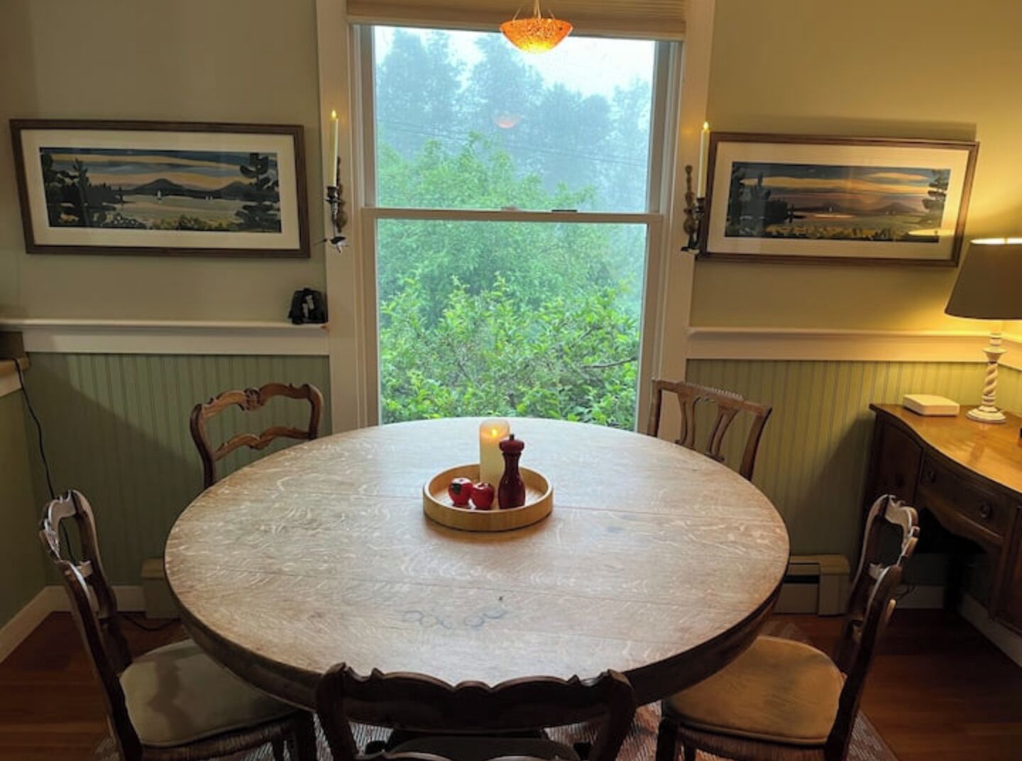Une table de salle à manger ronde, entourée de chaises, avec une bougie et un panier de fruits. Deux tableaux encadrés ornent les murs, une fenêtre offre une vue sur la verdure, et une lampe est posée sur une petite table en bois.