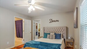 Quarto com cabeceira estofada, cama com colcha azul e branca, mesa de cabeceira com abajur e ventilador de teto.