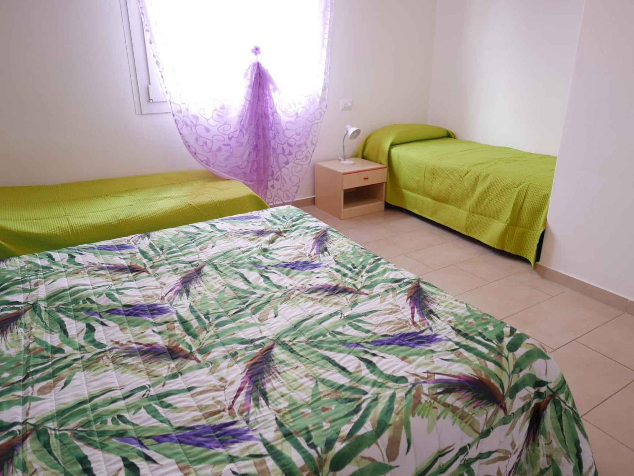Una stanza con due letti singoli, un piccolo comodino con una lampada e una finestra con tenda leggera.