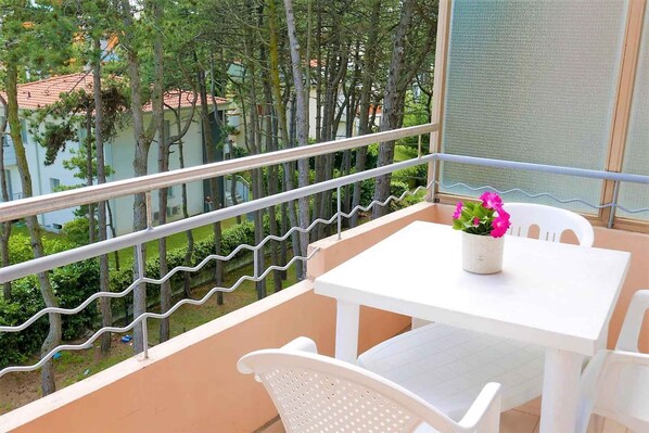 Un balcon avec une table et des chaises blanches, un pot de fleurs et une vue sur les arbres et les bâtiments.
