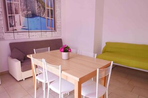 Un coin repas avec une table en bois et des chaises blanches, un canapé et un tableau représentant un paysage côtier.