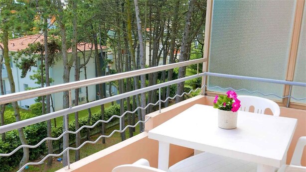 A balcony with a white table and chairs, a flower pot, and a view of trees and buildings. - Holiday apartment with air conditioning (San Michele al Tagliamento)