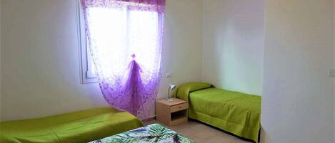 A room with two single beds, a small bedside table, and a window with purple curtains.
