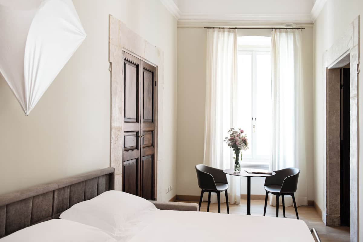 A hotel room with a bed, two chairs, a table with a vase, and a window with curtains.