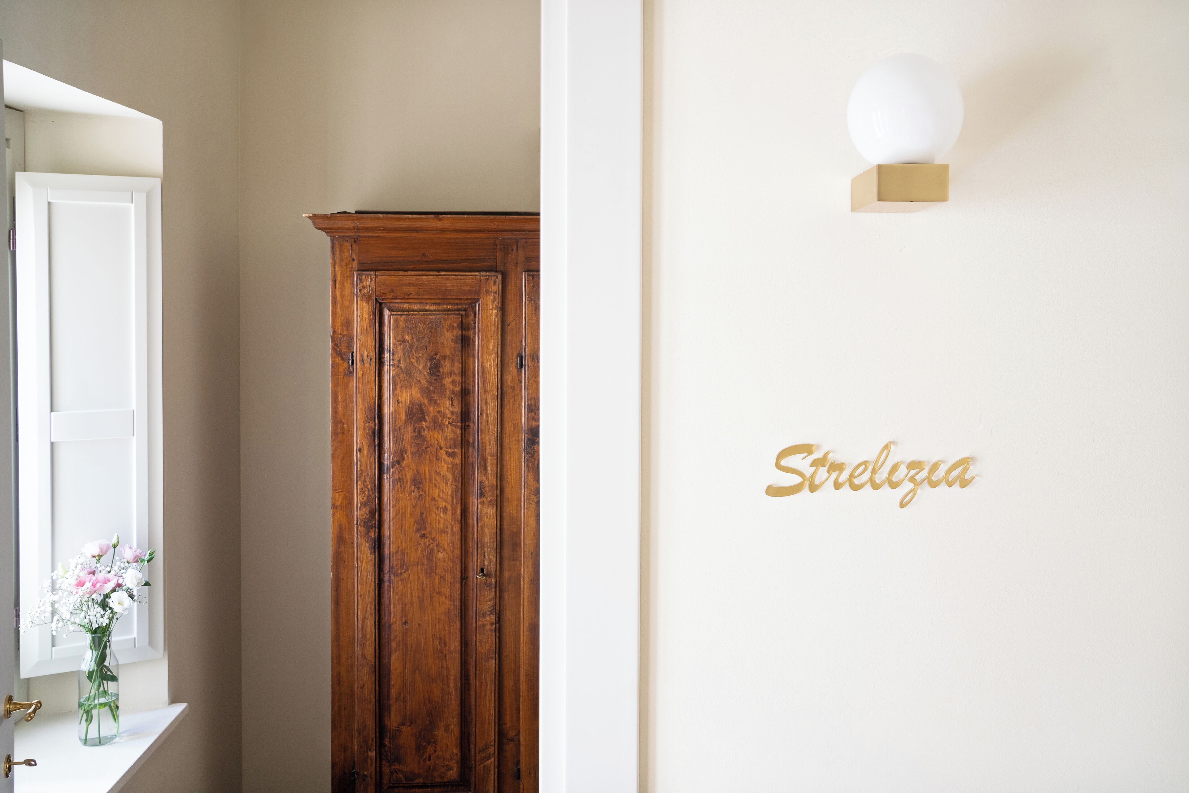 A hallway with a wooden wardrobe, a white wall with a gold sign, and a window with a vase of flowers.