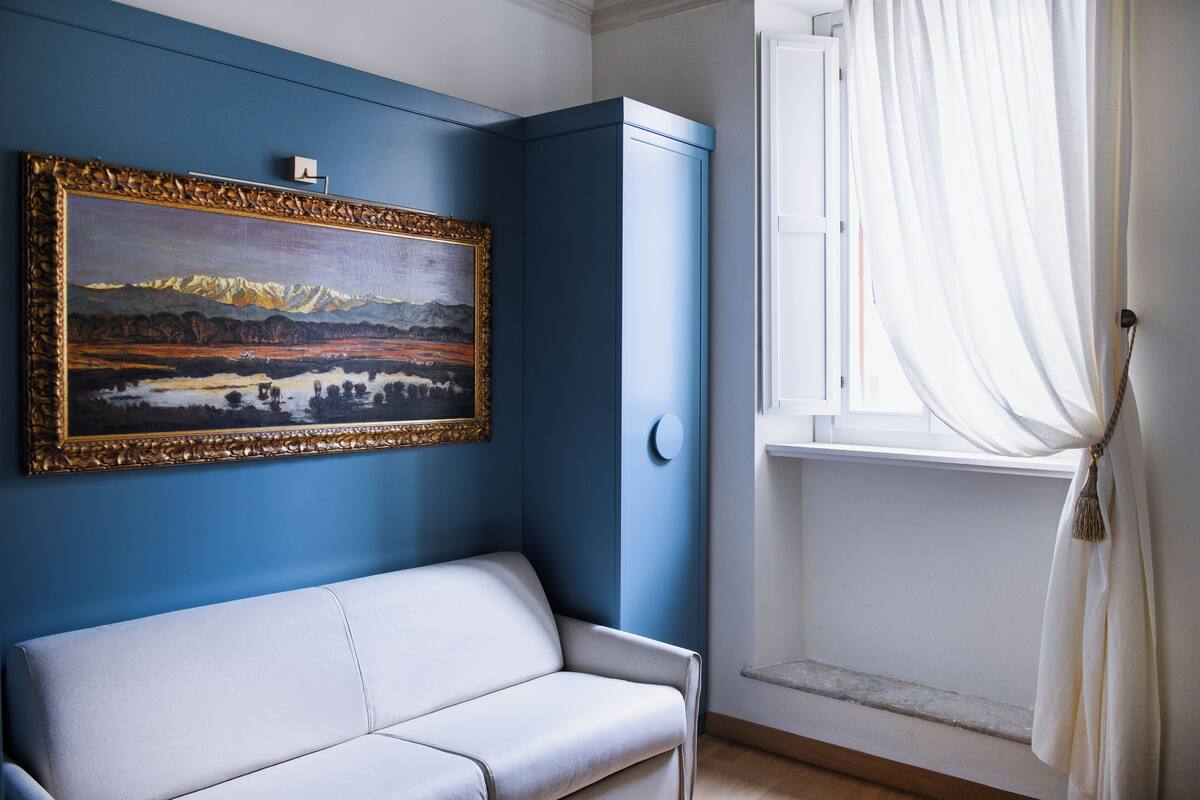 A room with a blue wall, a white sofa, a framed painting, and a window with curtains.