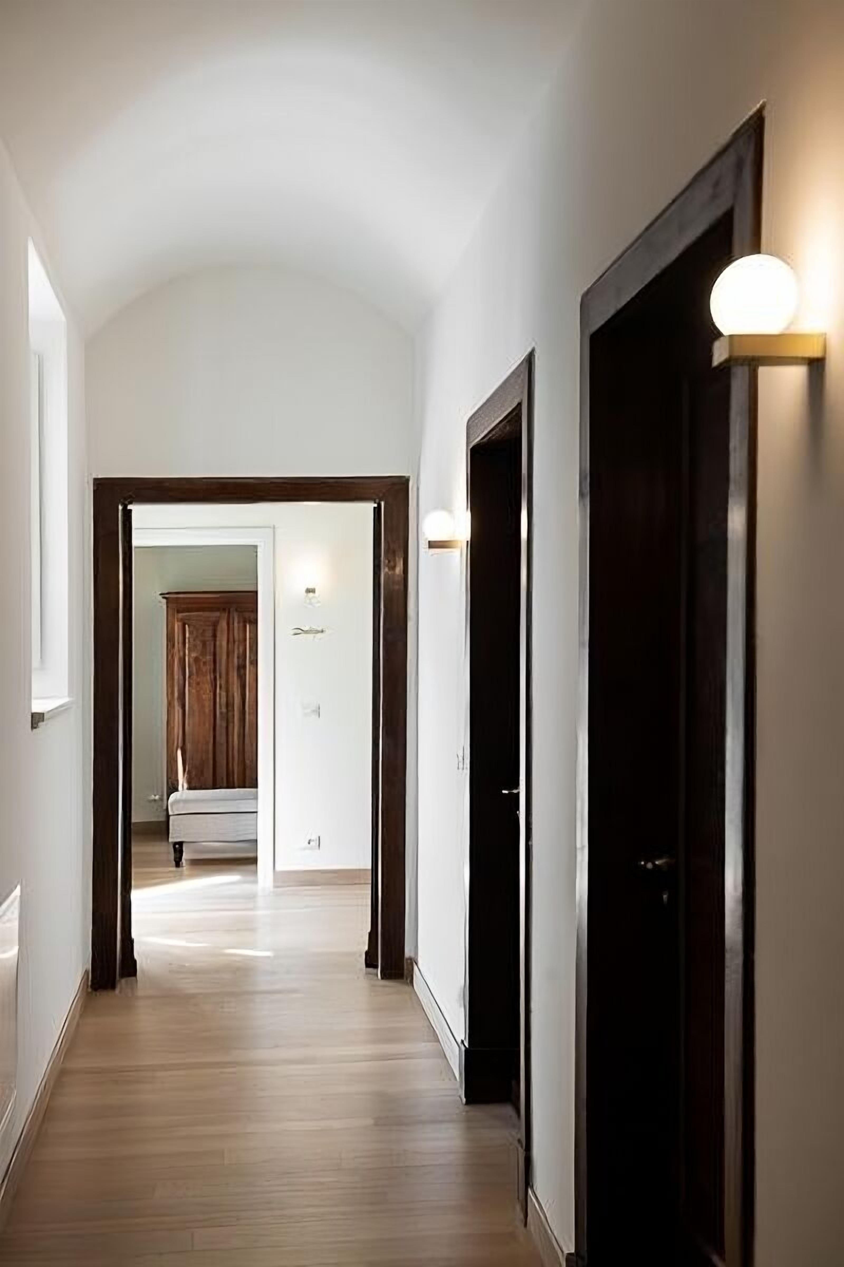 A hallway with wooden flooring, dark wooden doors, and wall-mounted light fixtures.