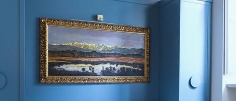 Quarto com parede azul de destaque, um quadro de paisagem emoldurado, sofá branco e piso de madeira.