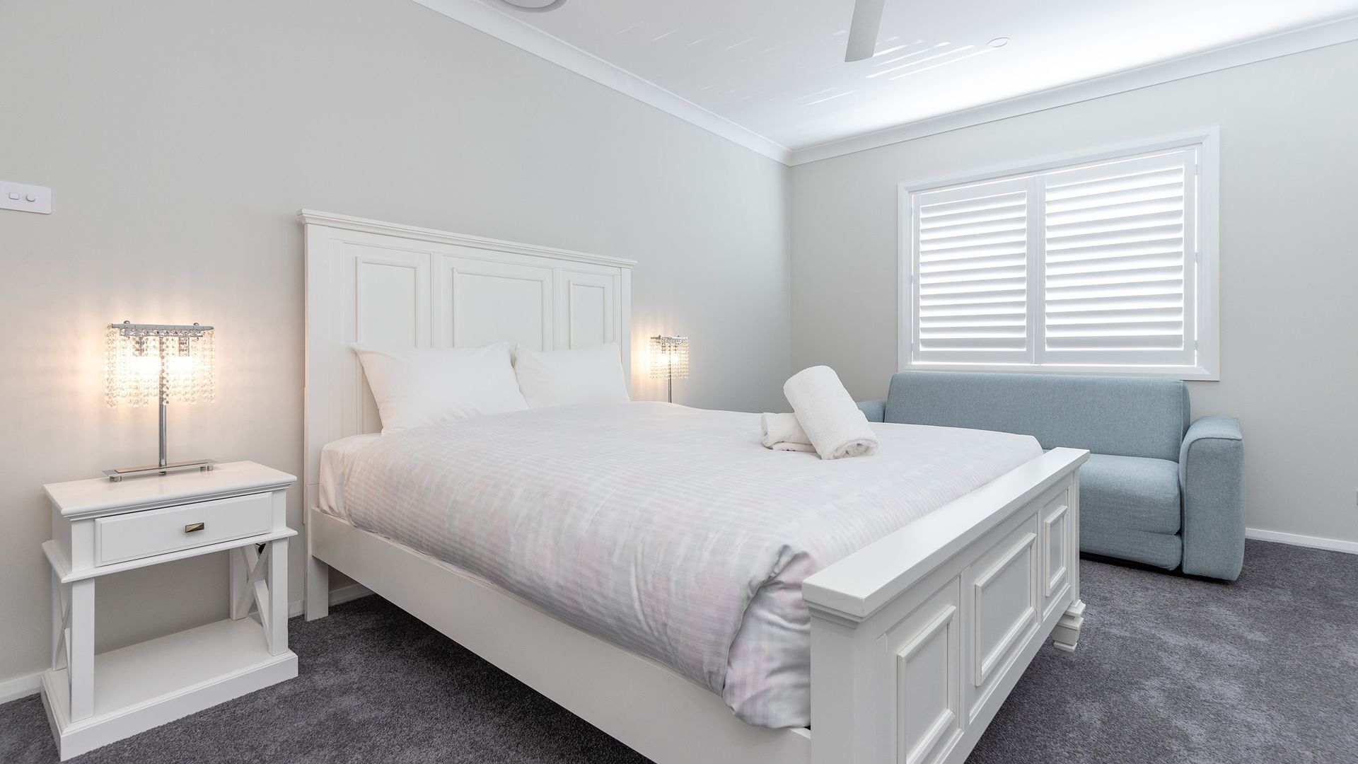 Un dormitorio con una cama grande, una mesita de noche, una lámpara de pie, una ventana con persianas y una silla azul.