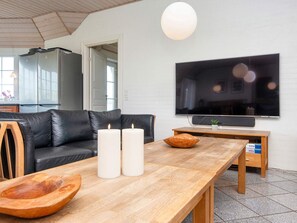 Un salon moderne avec un canapé en cuir noir, une table basse en bois, deux bougies blanches, un téléviseur à écran plat et une suspension.