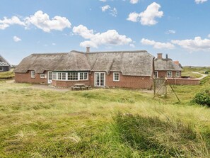 Et hus med stråtækt tag og murstensfacade, en træbænk og et gyngestativ i en græsplæne.