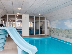 En indendørs swimmingpool med blå rutsjebane, glasvægge og et maleri af en strand.