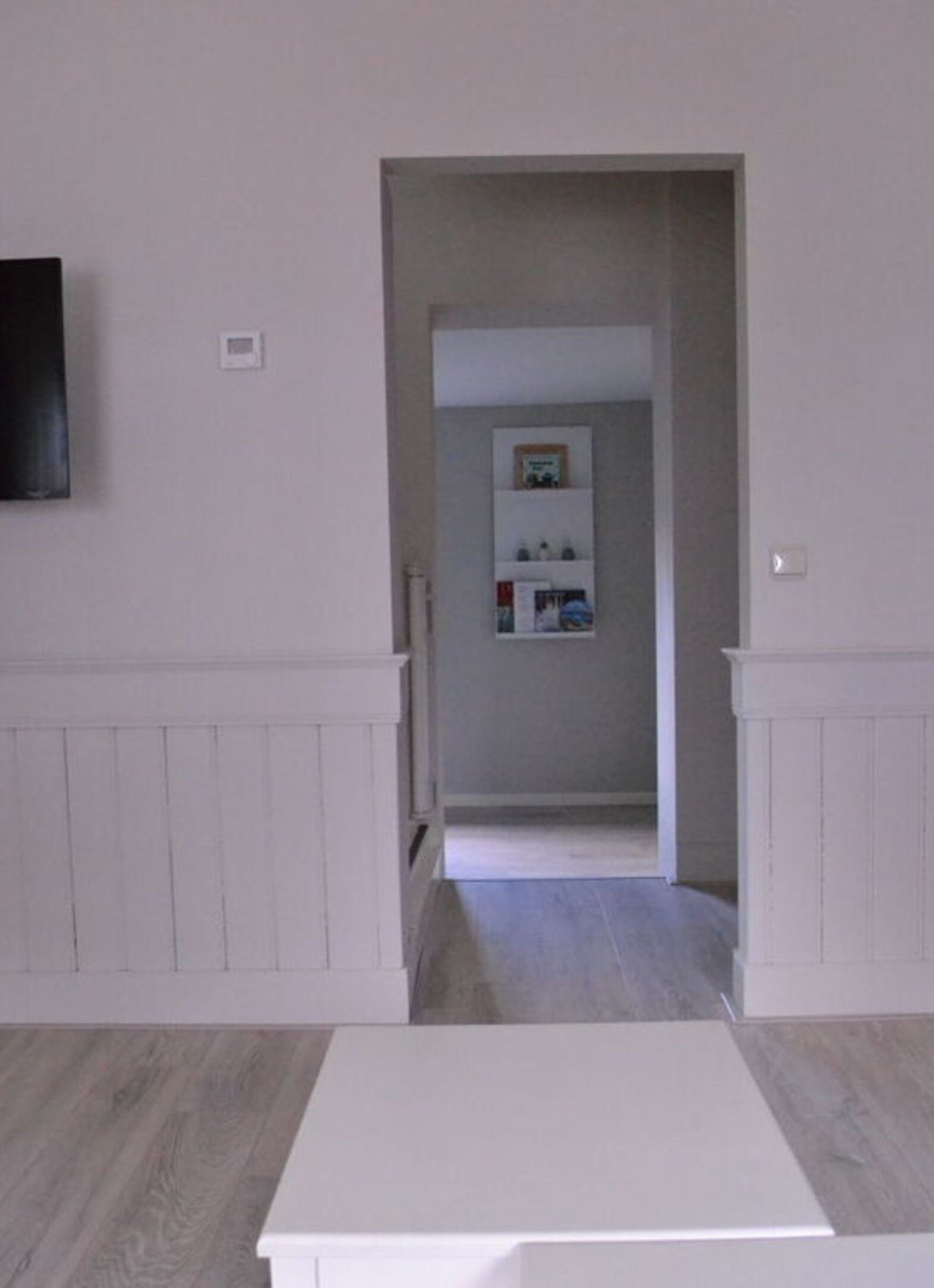 Een hal met een witte deur die leidt naar een kamer met een wit kastje en een klein boekenplankje.