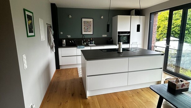 A modern kitchen with a central island, white cabinetry, and a large window providing a view of greenery outside. - Oasis of peace near the city center: Stylish garden apartment, 2 bedrooms, top public transport links  (Düsseldorf)