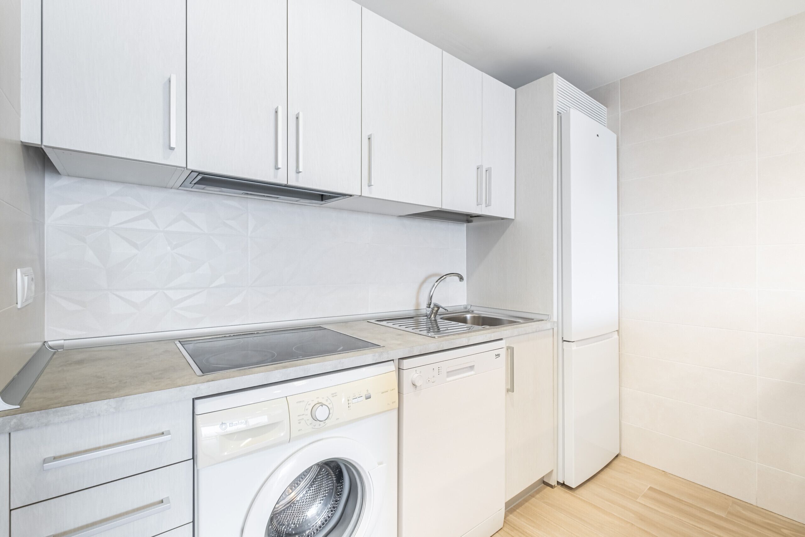 Cocina compacta con armarios blancos, lavavajillas integrado, lavadora y refrigerador.