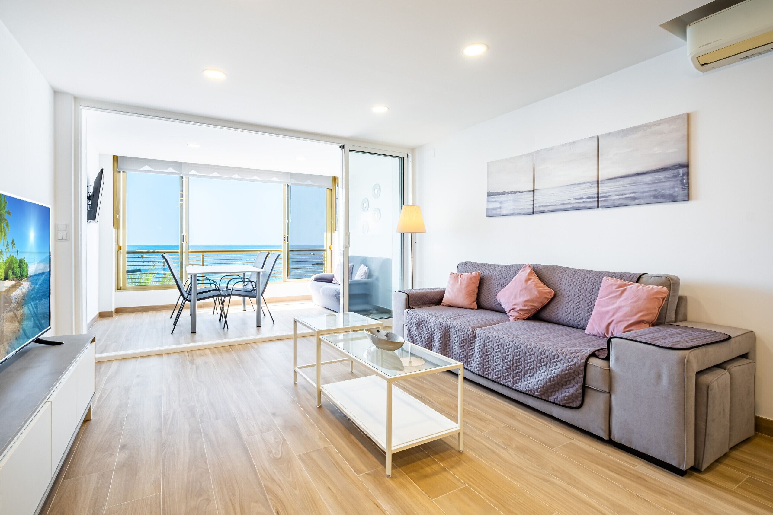 Una sala moderna con un sofá, una mesa de centro de vidrio y un televisor. Hay un balcón con vista al mar.