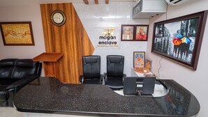 A hotel reception area with a marble wall, two black leather chairs, a wooden desk, and a clock. - Magan Enclave (Tarangambadi)