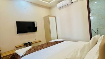 A hotel room with a bed, a flat-screen TV, a ceiling fan, an air conditioning unit, and a bedside table with a lamp.