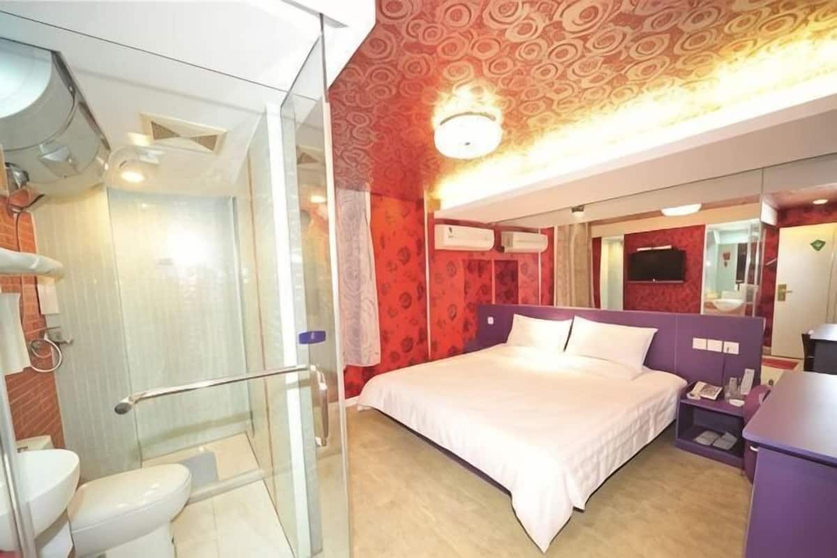 Hotellihuone, jossa on lasiseinäinen suihkutila, valkoisilla lakanoiden peitetty sänky, violetti pääty ja peili sängyn yläpuolella.