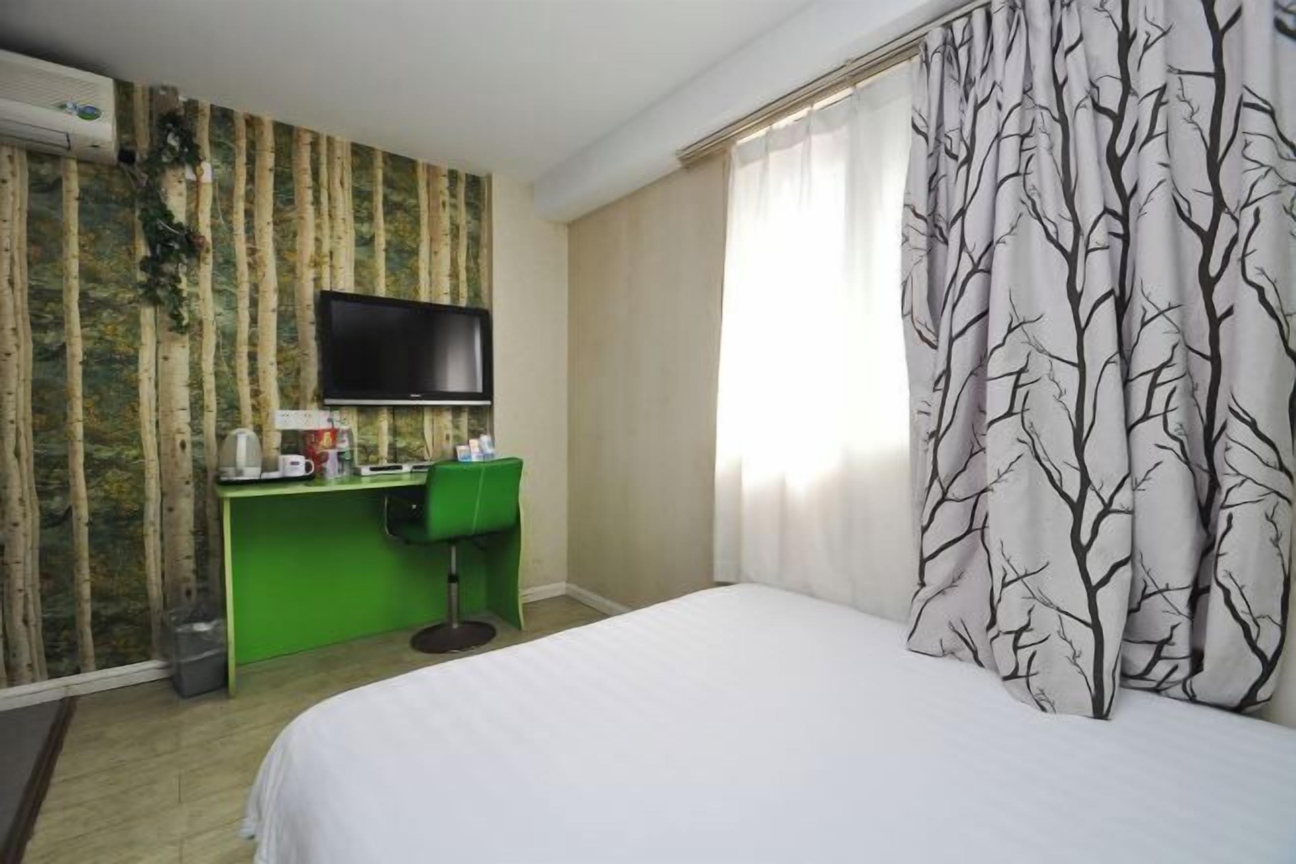 A hotel room with a bed, a desk with a flat-screen TV, a green chair, and a window with sheer curtains.