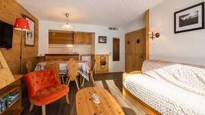 A cozy room with a bed, a red chair, a wooden table, and a kitchenette in the background. - 300m from ski slopes, 2nd floor, view mountain, balcony, fireplace or stove, tv, ski locker, 53m² (La Clusaz)