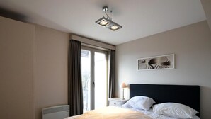 A modern bedroom with a bed, a bedside table, a radiator, and a window with curtains. - Golf en Strand Apartment Near the Beach (De Haan)