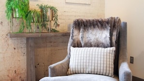 Un fauteuil gris, agrémenté d’un plaid en fourrure et d’un coussin à carreaux, est placé dans une pièce dont le sol est en bois et un des murs est en briques.