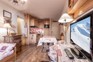 A cozy room with a wooden floor, a dining table with a patterned tablecloth, a television displaying a snowy mountain, and a kitchenette with wooden cabinets.